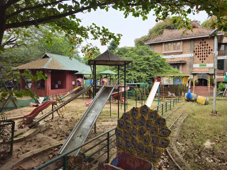 A children’s play area with slides and a painted peacock inside the zoo grounds, with tiled buildings and heavy tree cover behind