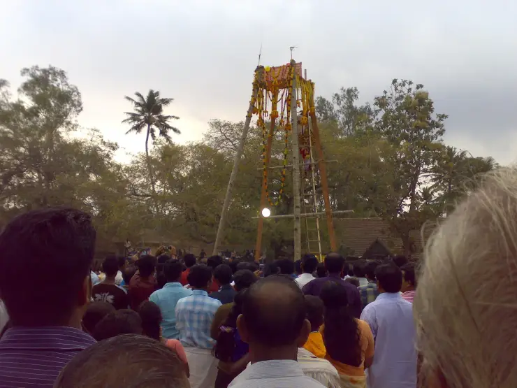 A tall garlanded wooden festival structure rising above a dense crowd at the Kaliyoot, Sarkaradevi Temple, Chirayinkeezhu