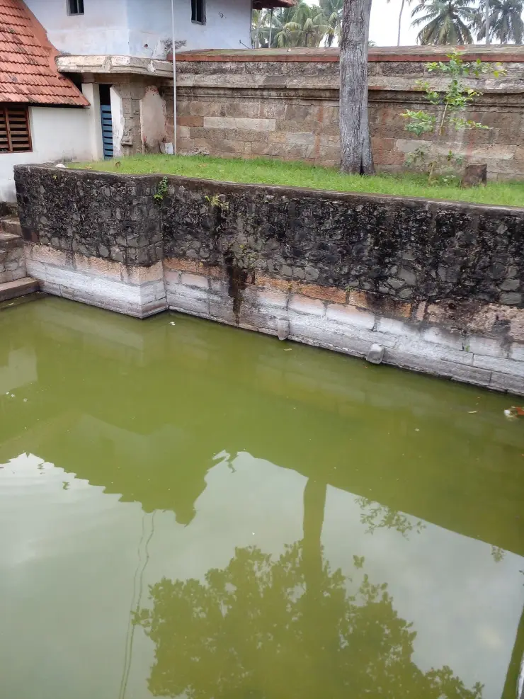The stone-lined temple tank at Thiruvallam, with laterite and granite walling and a tiled temple roof beyond