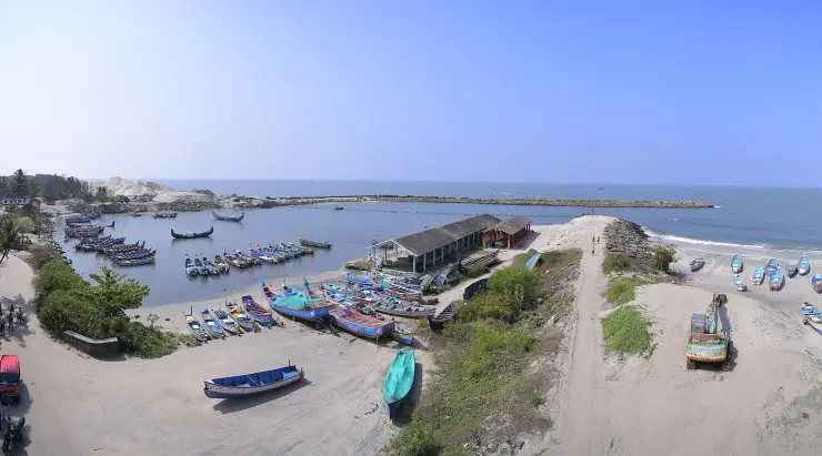 An elevated view of Thottappally harbour, its basin packed with painted fishing boats between two rock breakwaters, with heaps of dark sand and an excavator