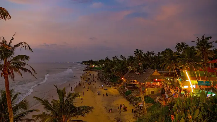 The lit café strip along the top of Varkala cliff at dusk, with rock armour piled at the base below