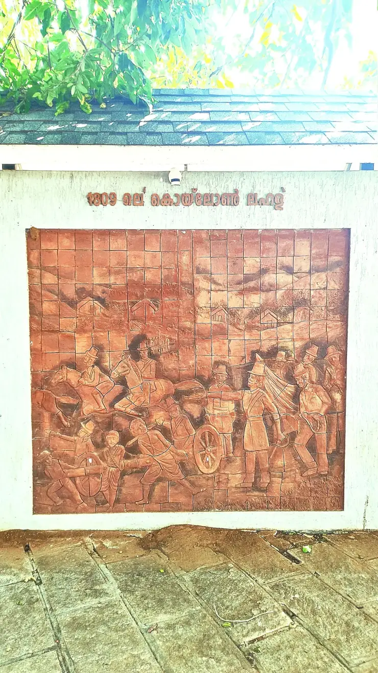 A terracotta relief panel set into a white wall under a tiled roof, captioned in Malayalam, showing cavalry, sepoys with muskets and a field gun being hauled by hand in a battle scene