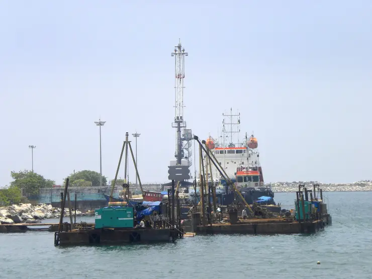 Pile-driving and dredging barges with a crawler crane working in the sheltered water at Kollam Port, a survey vessel and tug behind them and the rubble breakwater along the horizon