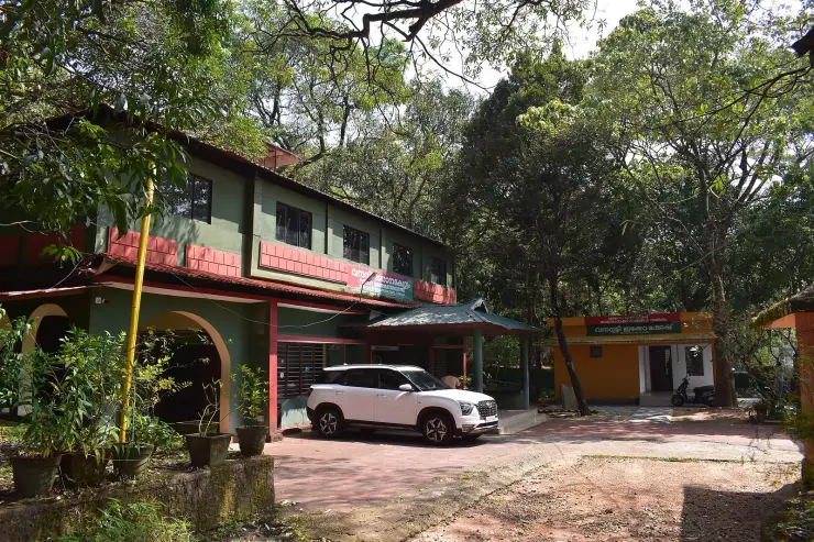 A two-storey green and dark-red Forest Department building with Malayalam signboards under a tiled roof, a car parked on the apron, and a small orange-walled eco shop beside it under tall trees.