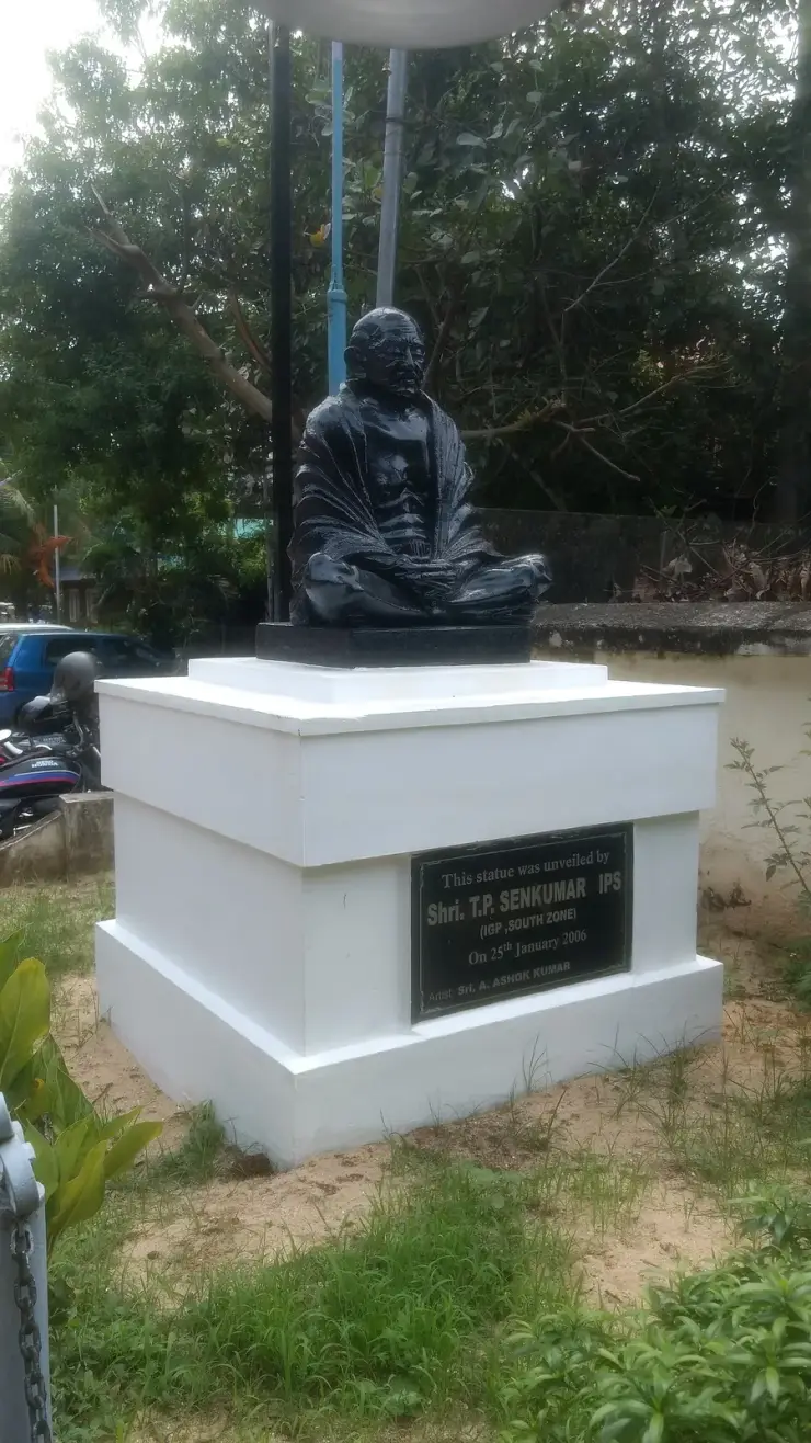 A black statue of a seated, cross-legged Gandhi on a white plinth in a garden, with a small dedication plaque on the front of the plinth