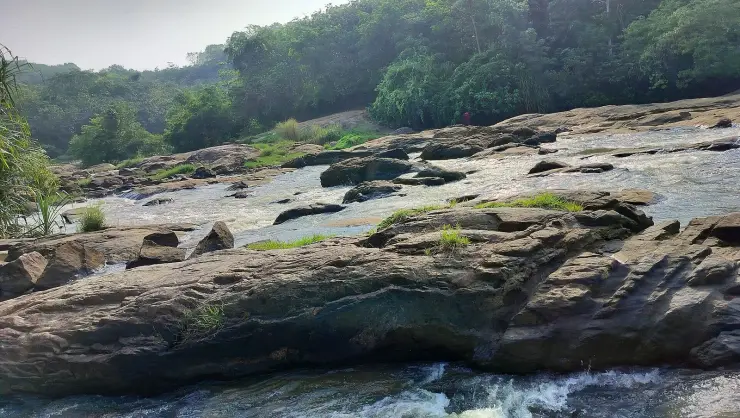 Long fluted ledges of grey rock stepping down into the river, with white water braiding between them and a single figure in red standing on the far bank for scale