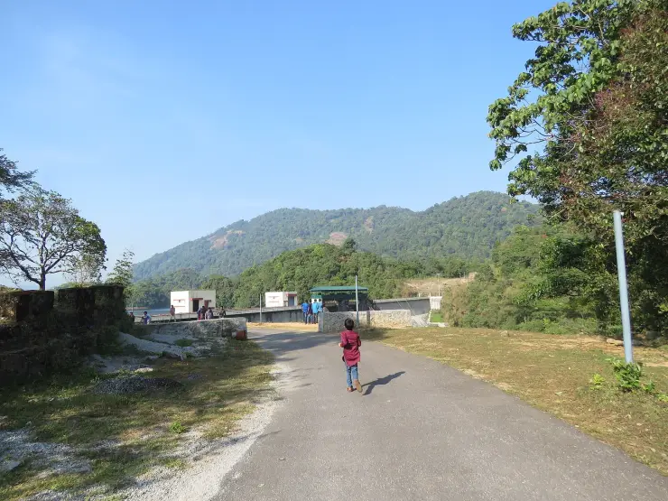 Visitors walking the metalled road along the crest of Kakkayam dam, past the white gate housings and a green-roofed shelter, reservoir to the left.