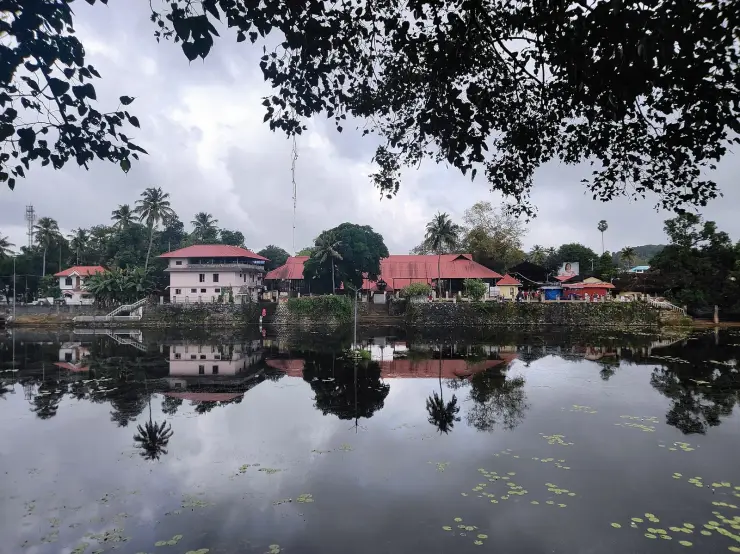 The Kottarakkara temple complex seen across its large rectangular tank at dusk, long red-tiled roofs and a white building reflected in still water with lily pads, laterite retaining walls dropping straight into the tank