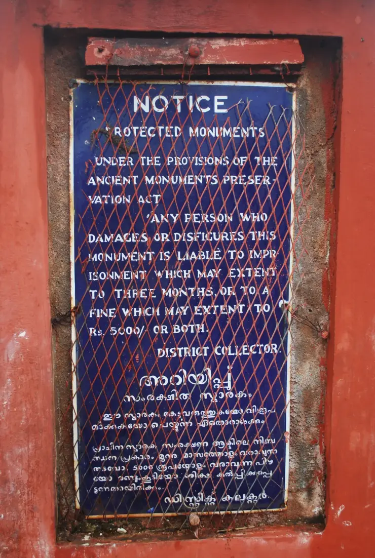 A blue enamel notice board behind rusted chicken wire reading NOTICE, PROTECTED MONUMENTS, under the provisions of the Ancient Monuments Preservation Act, signed District Collector, with the same text repeated in Malayalam.