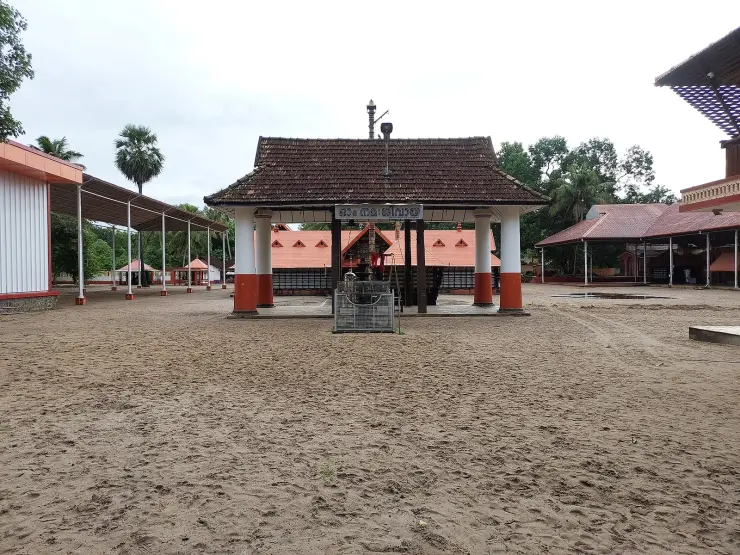 The sand courtyard of the Mararikulam Mahadeva temple, with a tiled namaskara mandapam, orange and white pillars and a board reading Om Namah Shivaya