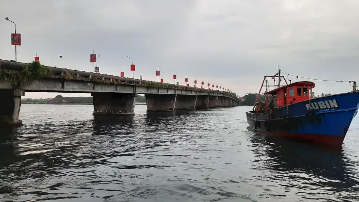 The low 1972 Neendakara road bridge on squat piers at water level, a large pipeline running along its flank and red banners on its lamp posts, with a blue-and-red trawler named SUBIN moored in the right foreground.