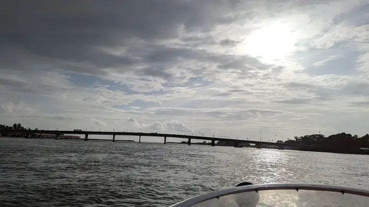 The high-level NH-66 bridge over the mouth of Ashtamudi Lake at Neendakara, seen low from a boat on the water, its long deck carried on tall piers with the auction hall visible at the far left of the shore.