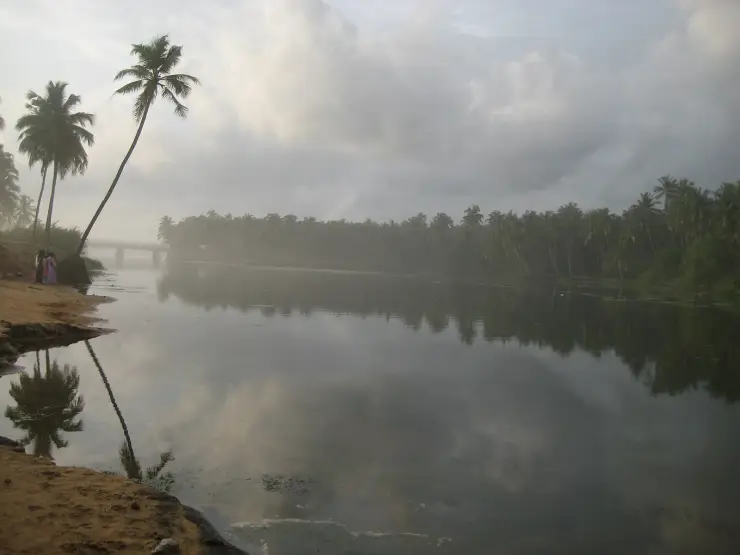 Still backwater at Paravur reflecting a cloudy sky, with a low bridge in the middle distance, a wall of coconut palms along the far bank, and a sandy spit in the foreground.