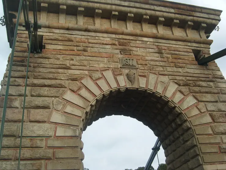 The masonry tower of the Punalur suspension bridge from below, showing a carved panel reading 1877 above the arch and a carved conch shell on the keystone beneath it.