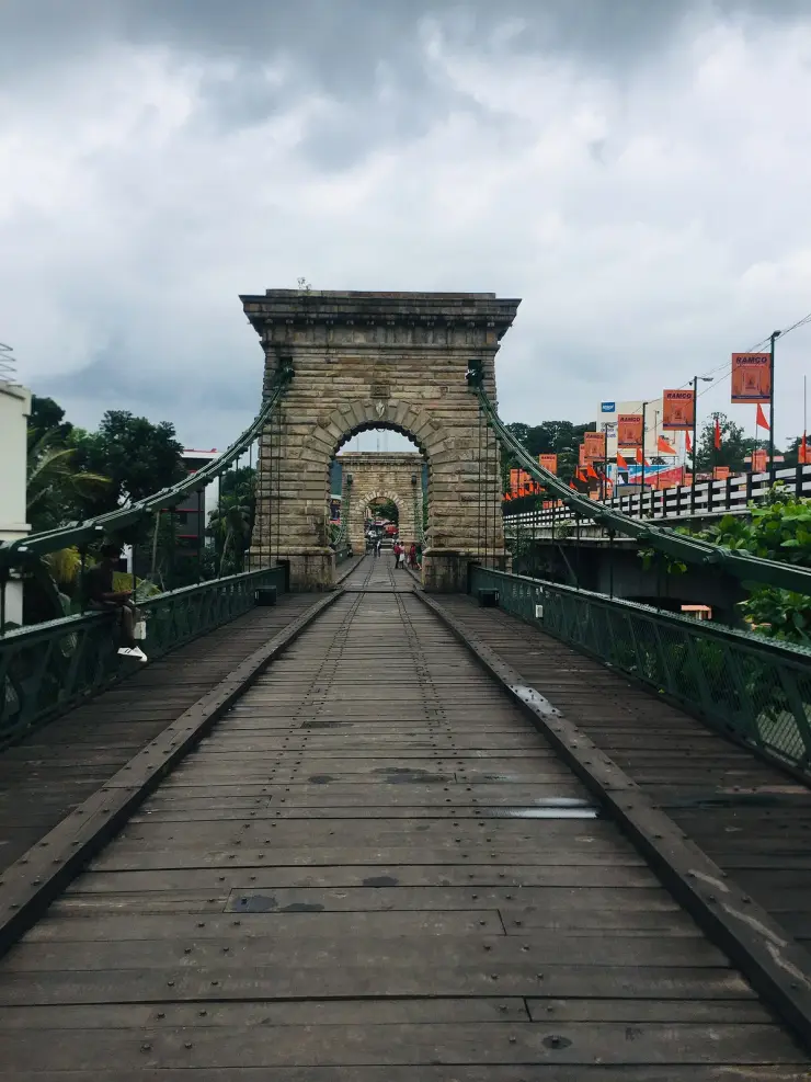 Looking down the timber deck of the Punalur suspension bridge under a storm sky, with two raised timber runners running the length of the planking towards the masonry arch.