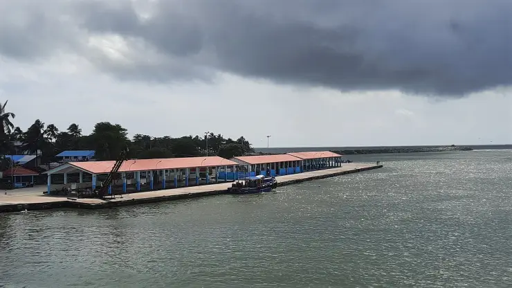 The long red-roofed open-sided auction hall on blue pillars along the Sakthikulangara quay, an ice-conveyor gantry at the near end and the breakwater running out to sea behind it with a light on its head, under a heavy grey storm sky, with a single small blue boat alongside an otherwise empty quay.