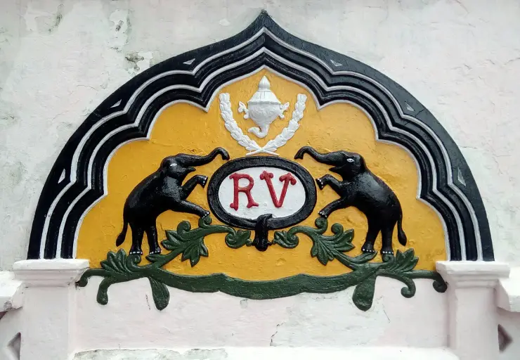 A moulded and painted crest set into the wall of Thevally Palace: a black many-lobed arch with white fillets around a yellow field, a white oval bearing the red monogram “RV” at the centre, two black elephants rearing with trunks raised on either side, a white conch-like device in a wreath above, and a green foliate scroll below.
