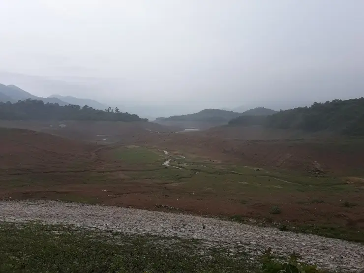 A wide reservoir basin drawn down to bare red earth, with a narrow stream channel winding through green flats on the valley floor and forested ridges disappearing into low monsoon cloud beyond.