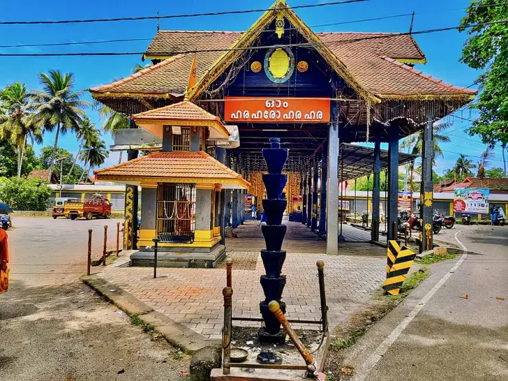 The street-front entrance of the Haripad temple with tiled roofs, an orange Malayalam board and a tall tiered black stone lamp pillar in the foreground