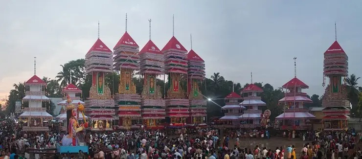 Eleven tall tiered festival edifices with red canopies and spire finials raised over a crowd of thousands in a field at dusk, with a giant painted standing figure at left