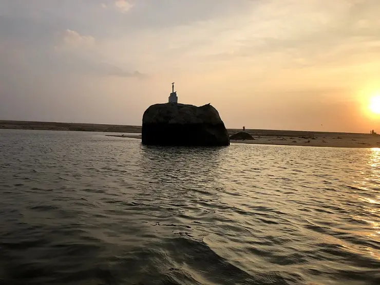 A large dark rock standing in the Poovar estuary at sunset, with the sandbar and the open sea behind it