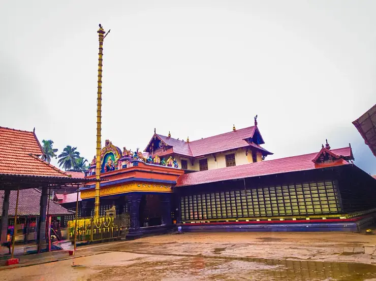 The gilded flagstaff of the Haripad Subrahmanya Swamy temple rising against tiled roofs and a painted gopuram, courtyard wet with rain