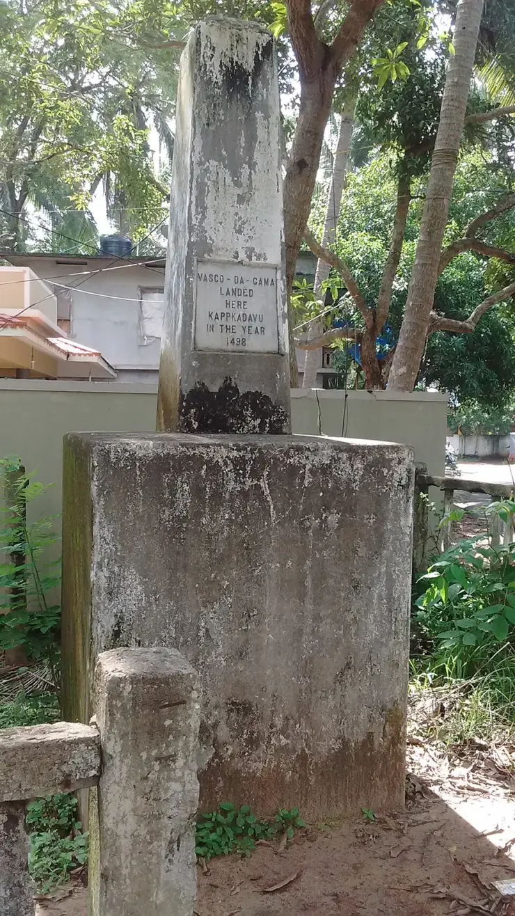 The weathered stone Vasco da Gama pillar at Kappad on its square plinth, its white inscribed tablet reading “Vasco-da-Gama landed here Kappkadavu in the year 1498”, with houses and coconut palms behind.