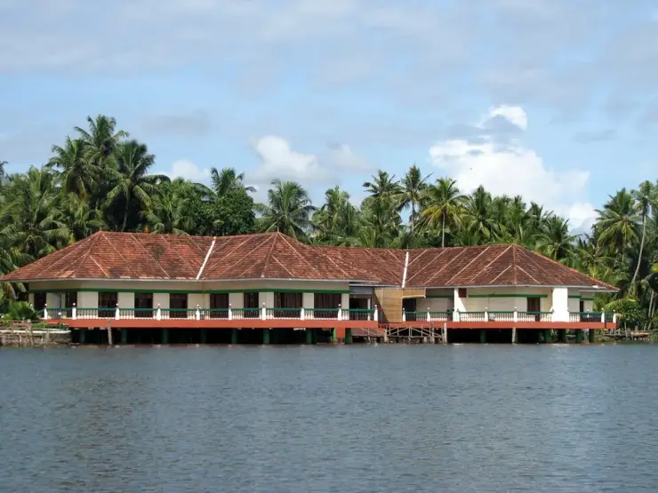 A long low tile-roofed building standing on green and white posts directly over calm backwater, palms crowding behind it