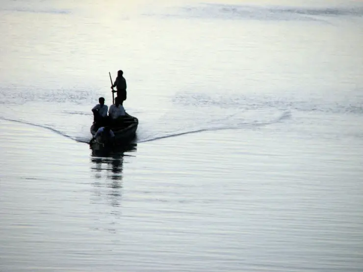 A country boat in silhouette on wide pale water at dusk, three figures aboard, one standing with a pole, wake spreading behind