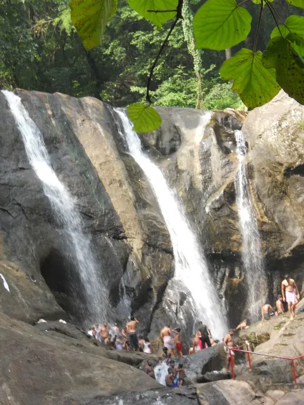 Dozens of people standing on a rock shelf directly beneath four narrow chutes of falling water, with red steel railings to one side and dark hollows scoured into the cliff.