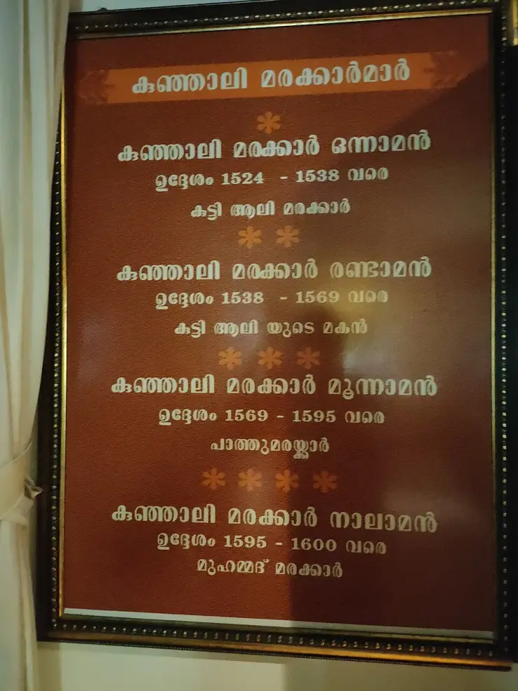 A framed Malayalam board hanging in the Kunjali Marakkar Museum, headed “കുഞ്ഞാലി മരക്കാർമാർ”, listing the four Kunjali Marakkars with their names and dates, each date preceded by the word “ഉദ്ദേശം” — approximately.