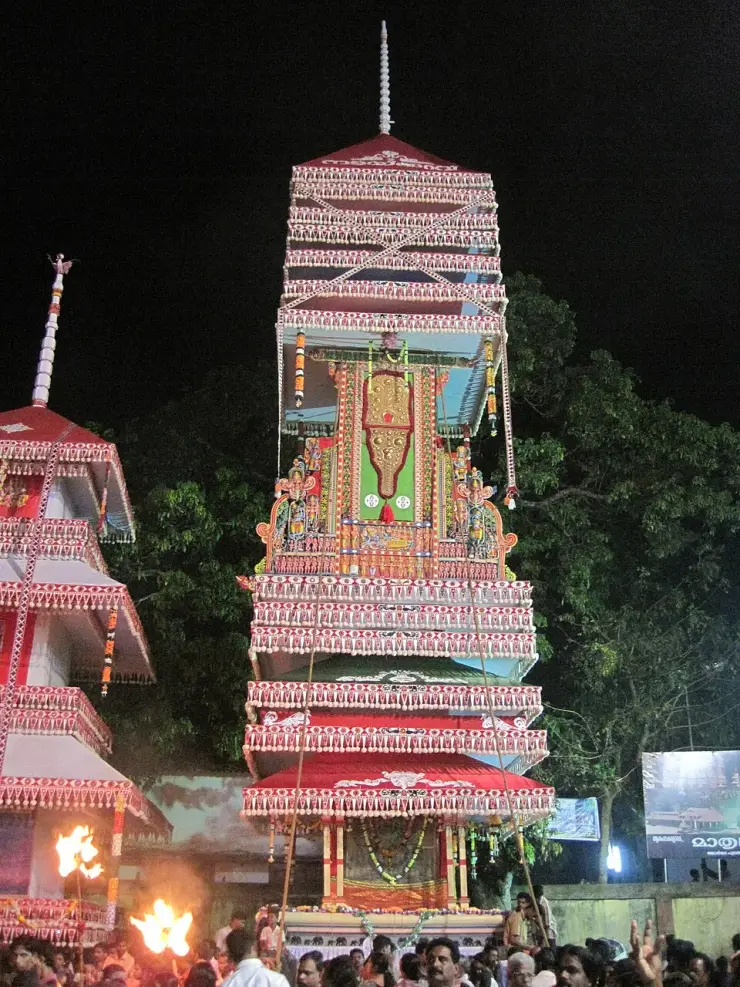 A single tall tiered Kuthira tower lit by torches at night, hung with tinsel fringes, a painted central panel and a spire finial