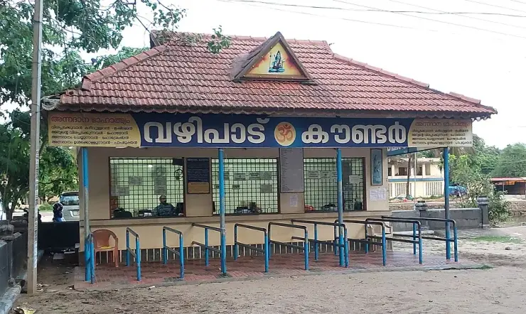 The vazhipadu offerings counter at Oachira temple — a shuttered booth with printed rate boards listing offerings and their prices.