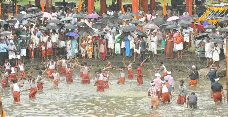 Men in orange mundus and white vests standing thigh-deep in a flooded field, sticks raised above their heads, while a crowd watches from the bank under black umbrellas in heavy monsoon rain.