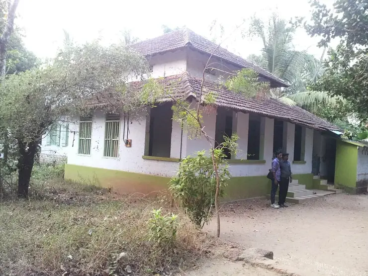 Puzhakkara Palli at Chaliyam: a low whitewashed mosque with a green-painted plinth under a two-tiered clay-tiled hipped roof, wooden pillars along an open verandah, two men standing on the step, a red-earth lane and thick greenery around it.