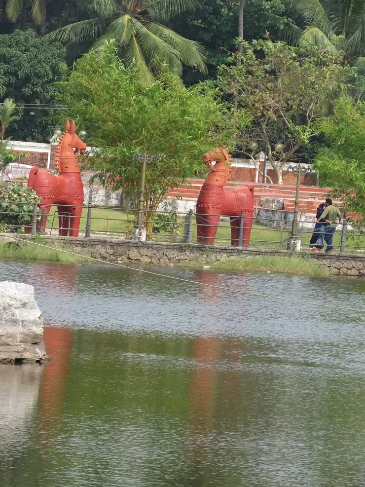 Two large red terracotta horses standing on the bank above the quarry lake at Sargaalaya, Iringal, with visitors walking the path behind them