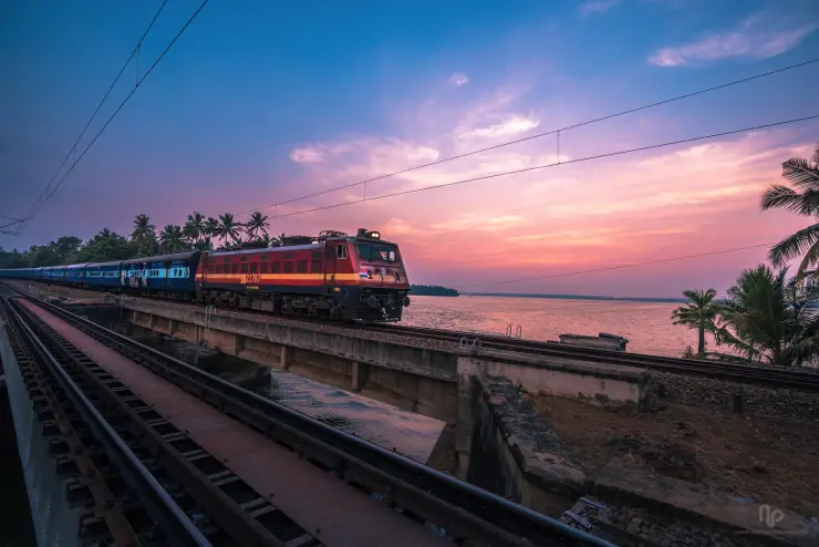 A red electric locomotive hauling blue passenger coaches across Peruman Bridge beside Ashtamudi Lake at sunset.
