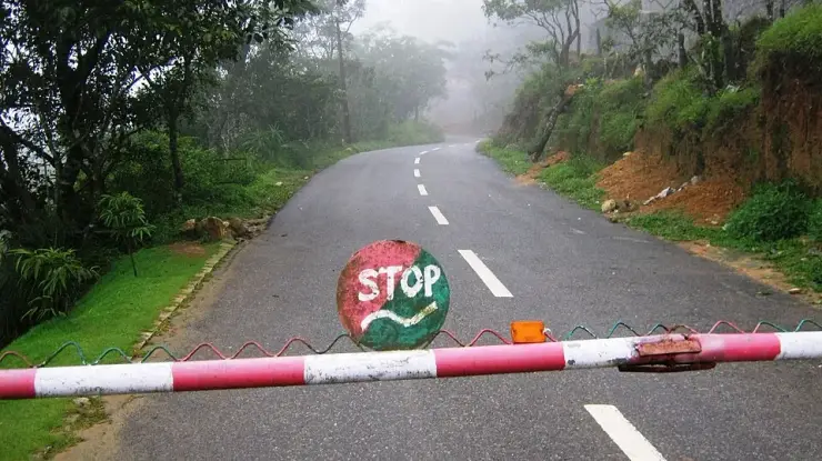 A red and white barrier lowered across the misty Ponmudi ghat road at the forest check-post, with a hand-painted stop sign