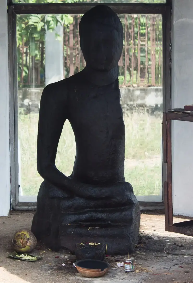 The Karumadikuttan Buddha with its left arm missing, and on the floor before it a split coconut on a banana leaf, loose flowers, a brass bowl and a stone lamp