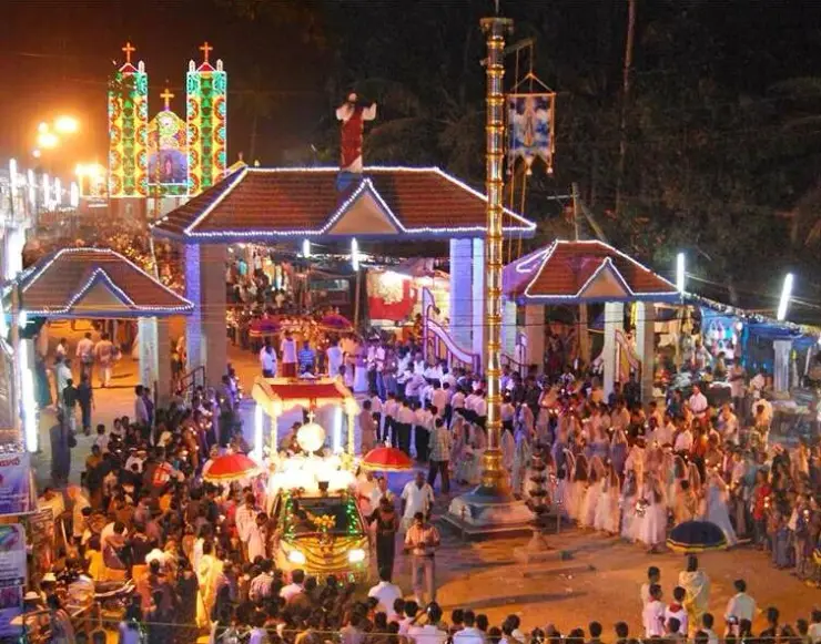 A night procession fills Pallikkunnu’s decorated gateway, with an illuminated church and golden flagstaff behind the crowd.