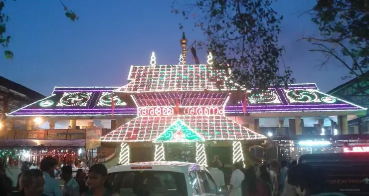 The Malayalapuzha temple entrance is illuminated at night, with a crowd gathered on the approach.