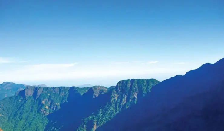 Sunlit cliffs and forested slopes rise beneath a blue sky in the Vellarimala range.