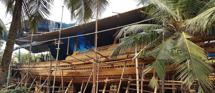 A large wooden uru under construction at Beypore, its pale planked hull propped on timber scaffolding beneath a woven roof, coconut fronds across the foreground.
