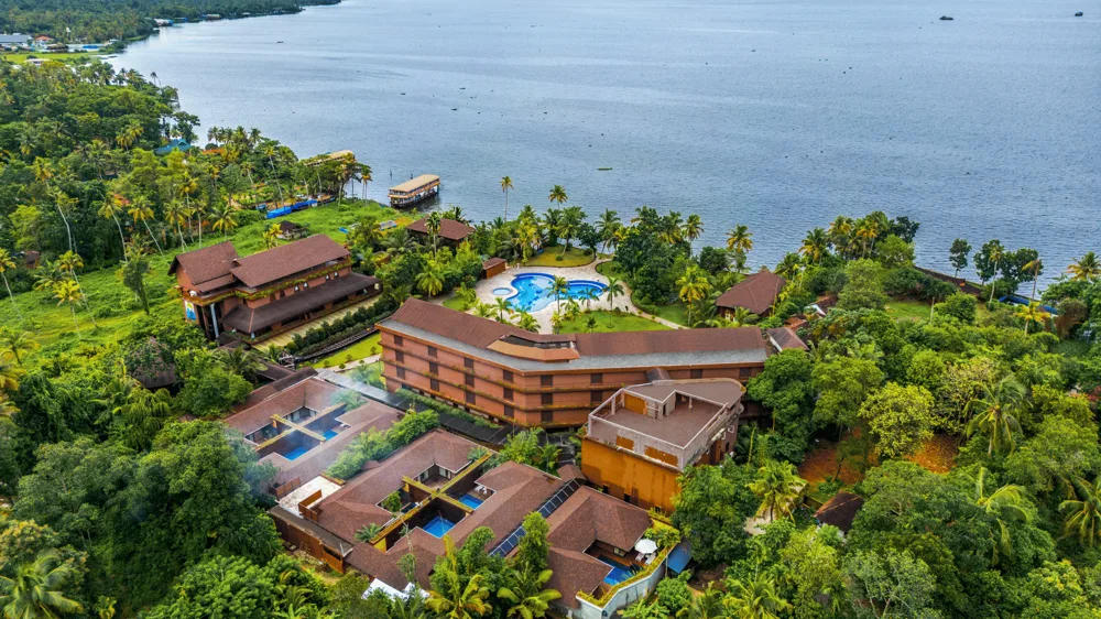 An aerial view of a hotel nestled among lush greenery, with a breathtaking expanse of the vast ocean and clear sky in the background - Uday Backwater Resort
