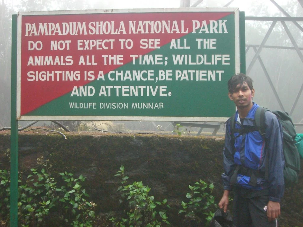 A visitor stands beside a forest sign in the Pampadum Shola landscape.