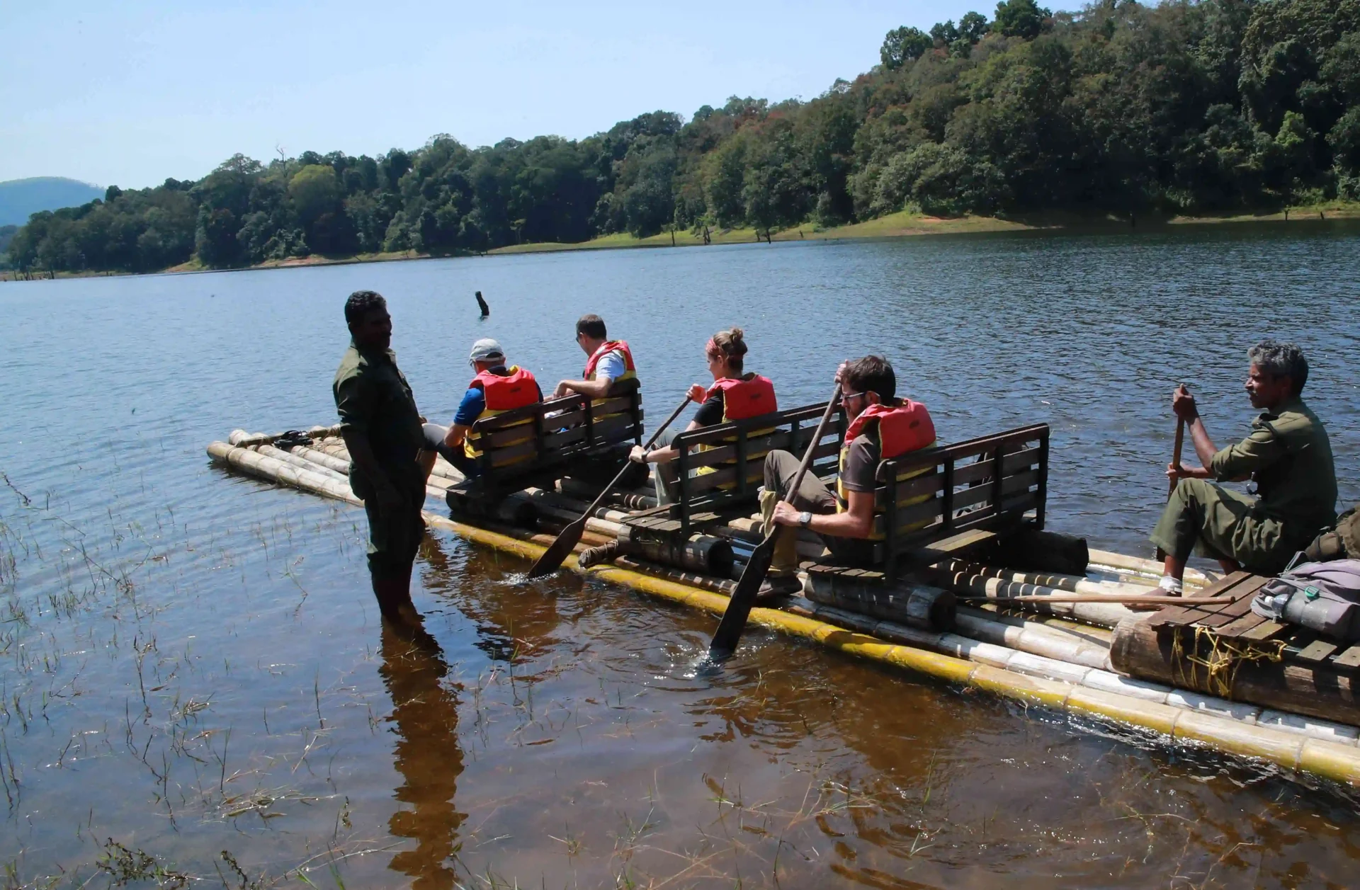 A guide stands beside a bamboo raft in shallow water while life-jacketed passengers sit in rows.