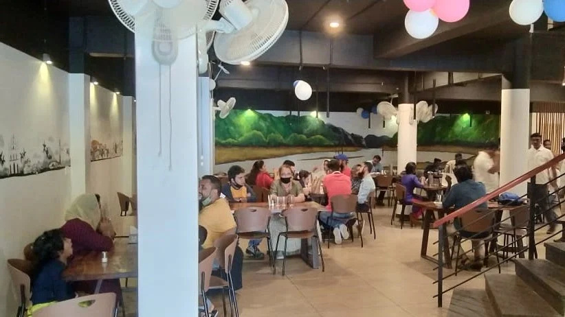 Tables, seated diners and a forest mural inside Periyar Tiger Cafe, with stairs at the right.