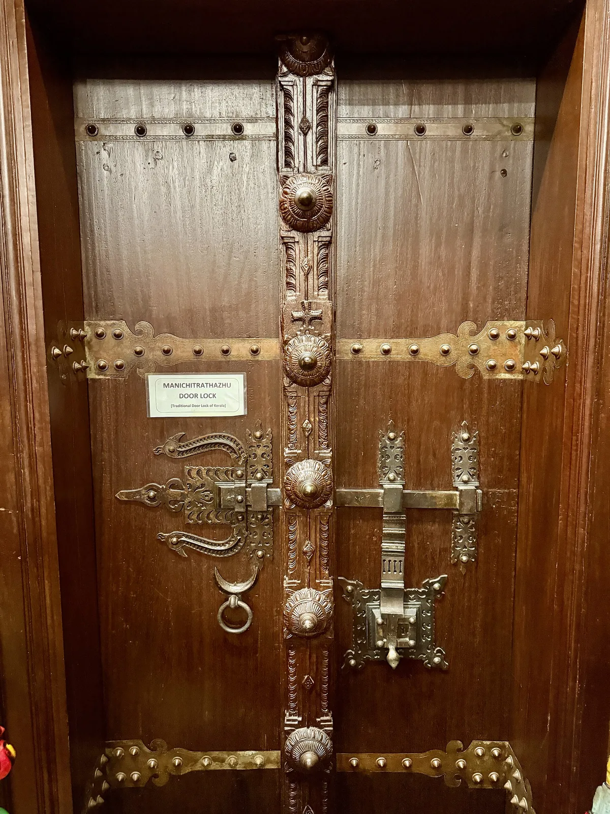 A carved teak door at the Kerala Folklore Museum fitted with an ornate brass manichitrathazhu lock, bolts and studded straps