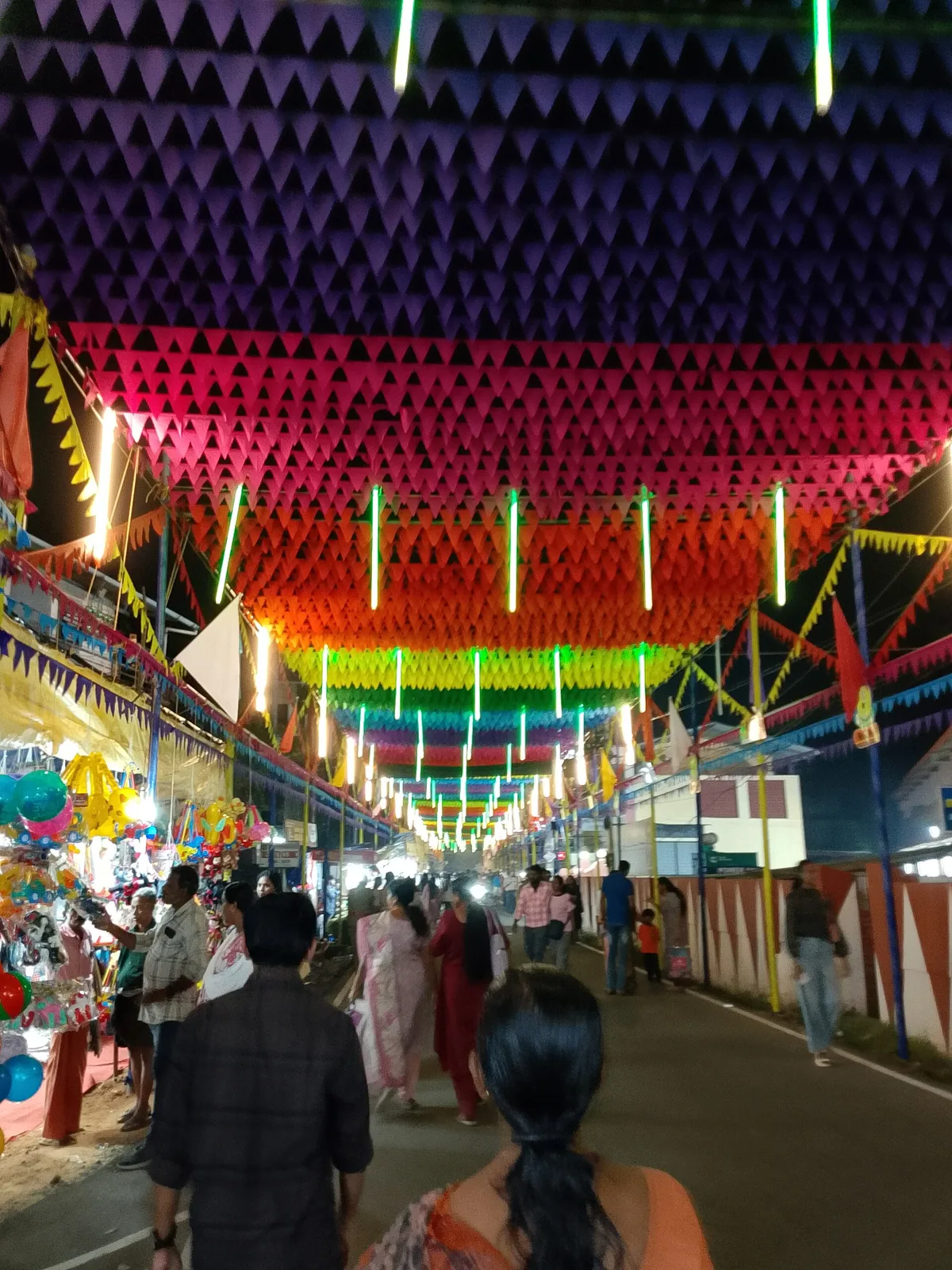 A street in Alappuzha roofed over with brightly coloured pleated decorations and strings of lights during Mullakkal Chirappu, crowded with people and stalls