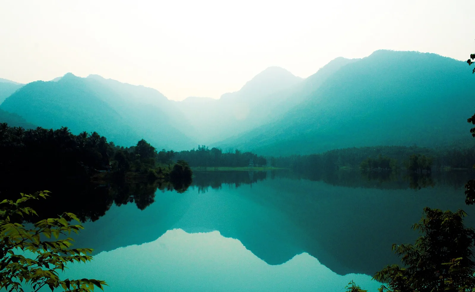 A still sheet of water at dawn seen from Kariyathumpara, with layered wooded ridges rising behind it in mist and the whole range reflected almost perfectly in the flat surface.